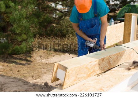 Construction worker or carpenter drilling a beam with wooden panels and interior insulation as he stands in the shade of a tree on a building site