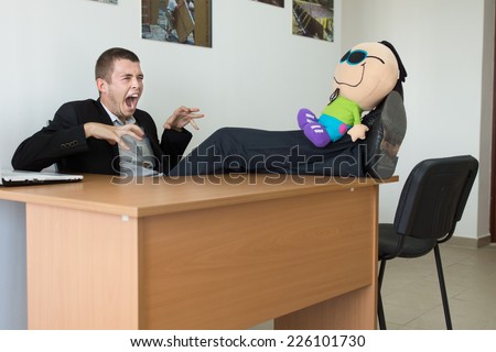 Young Male Office Worker Playing with Stuffed Toy, with Feet on the Table, Showing Monster Face at Office.
