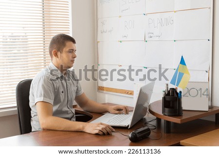 Young White Male Firm Manager Sitting at His Table Area Using His White Laptop Computer to Check Work Schedules.