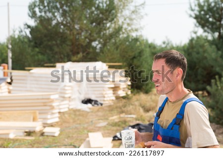 Young builder enjoying a coffee break standing laughing with a mug in his hand in front of stacked components for a prefab wooden house