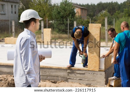 Young architect with a rolled blueprint under his arm standing watching construction workers on a building site