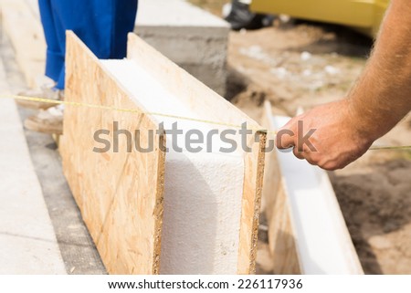 Workman measuring a prefab wooden wall panel with insulation on a construction site prior to installing it