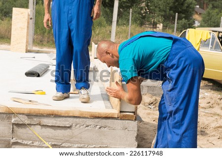 Construction worker in colorful blue overalls lining up insulated wooden wall panels on the foundation of a new build house watched by a co-worker