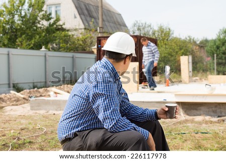 Builder on coffee break sitting on the construction site watching his colleagues continue working