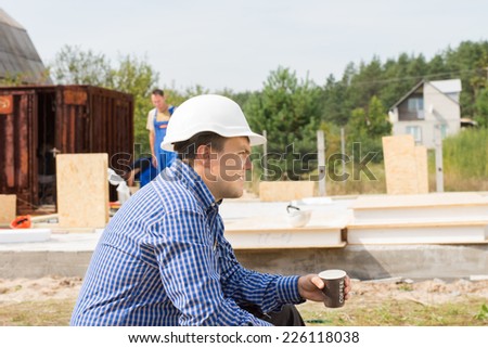 Thirsty builder taking a coffee break sitting side view with a mug of coffee in his hands as his crew continue work on the building behind him