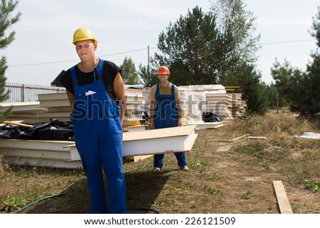 Two young construction workers in hardhats and overalls carrying a wall insulation panel on a building site