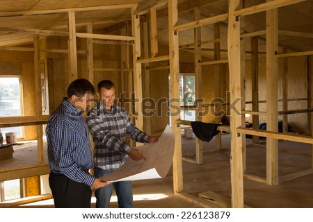 Two builders standing with an open blueprint discussing the interior of a half completed timber frame house