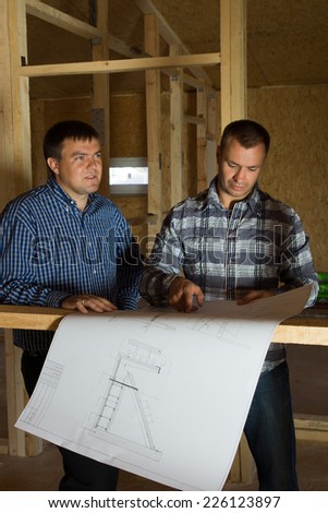 Two builders checking a blueprint together as they standing inside a half constructed timber frame house