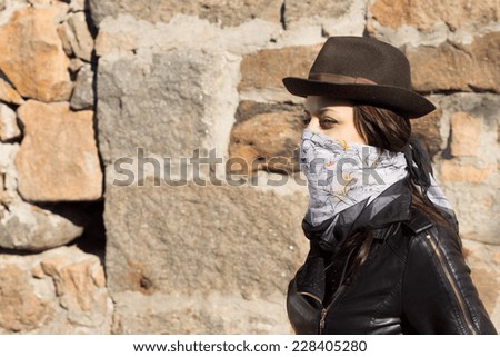 Young woman in bandit style fashion wearing a bandanna tied around her lower face in a trendy hat and leather jacket, side view in front of a stone wall