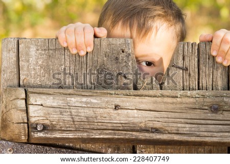 Little boy playing peek a boo through a gap in a broken plank in a rustic wooden gate looking at the camera with one eye