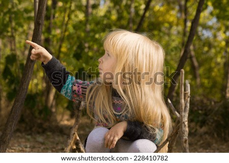 Little blond girl sitting on the ground in the garden pointing to the side and looking in the same direction