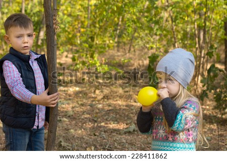 Cute little blond girl blowing up a colorful yellow party balloon outdoors in the garden watched by a young boy holding onto a tree trunk