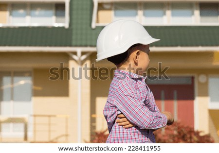 Close up Little Male Engineer Kid in White Helmet Looking at the House on the Side Seriously While Arms are Crossed.