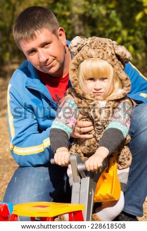 Cute little girl in a cat suit with spotted print posing in the arms of her father outdoors in the garden holding a colorful toy
