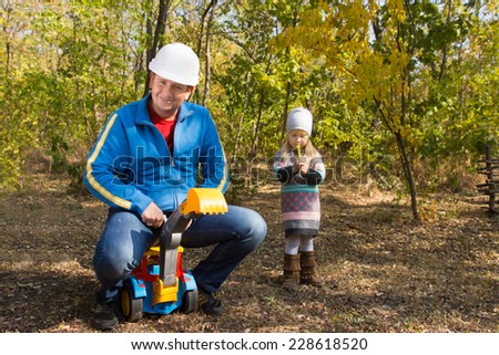 Father wearing a hardhat riding on his childs colorful plastic toy front end loader watched anxiously by a cute anxious little girl with her hands clasped