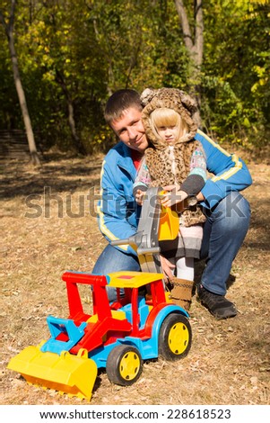 Cute little girl in a spotted cat suit posing in the arms of her kneeling father outdoors in q park with a large colorful plastic construction toy with a front end loader