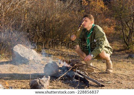 Hungry young scout cooking sausages over a small open fire in a clearing as he explores the wilderness