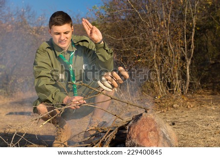 Scout saluting his cooking sausages as he bends down over a small camp fire in a clearing with the uncooked sausages speared on a twig