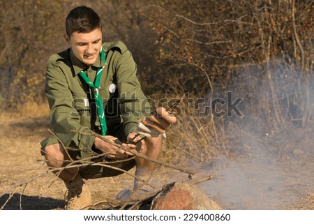 Young scout cooking his lunch over a fire as he squats down with a batch of sausages skewered on a twig with a smile of anticipation