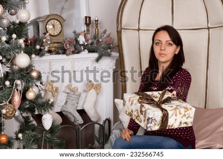Gorgeous Young Woman with Neutral Facial Expression in Offering Christmas Gift Sign Near Chimney Wall and Decorated Christmas Tree.