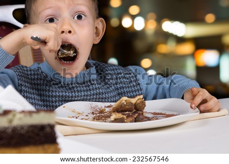 Hungry little boy gobbling down a slice of tasty chocolate cake with his mouth open wide for a mouthful as he sits at a table in a restaurant