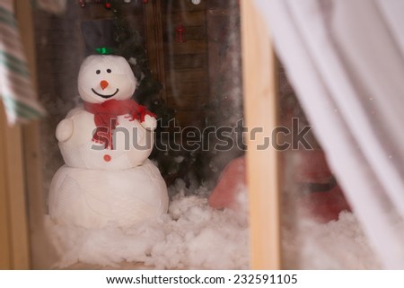 Christmas snowman viewed through a frosted window with open curtain standing in the winter snow outside in the darkness