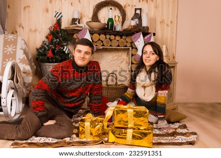 Young couple celebrating Christmas in a rustic cabin sitting on the floor on a rug in front of the hearth with stylish gold wrapped gifts smiling at the camera