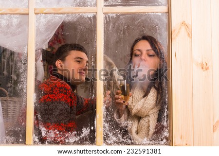 View through the cold frosted window of a young couple celebrating in a winter cabin enjoying a drink and blowing on the glass to condense their breath in the cold