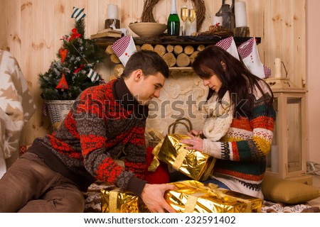 Young man and woman sitting on the floor in a rustic wooden cabin opening elegant metallic gold Christmas gifts eagerly untying the ribbon and bow