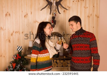 Happy Young Couple in Colored Winter Outfits Tossing Champagne Flute Glasses Inside the Wooden House for Christmas Celebration.