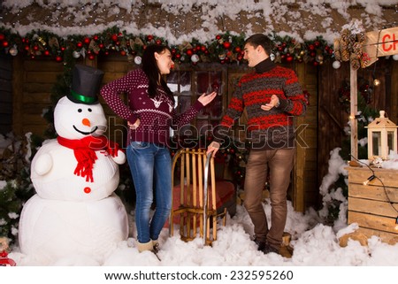 Young Couple in Winter Outfits Talking Inside the Wooden House, with Various Attractive Christmas Decors, Beside Big Indoor Snowman.