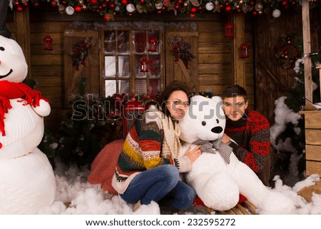 Sitting Young Couple in Winter Outfits Hugging White Winter Bear Doll at Wooden House Decorated with Big Snowman and Various Christmas Decorations.