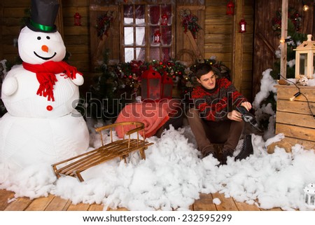 Young Handsome Man Sitting on Cotton Snow Decor on the Floor while Holding Ice Skates at Decorated Wooden House with large Winter Snowman.