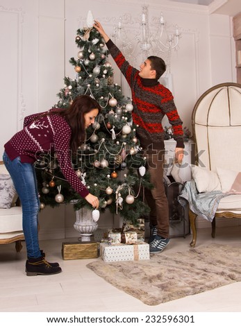 Young couple decorating a traditional green Christmas tree in a living room interior hanging decorations and stretching to place an ornament on top