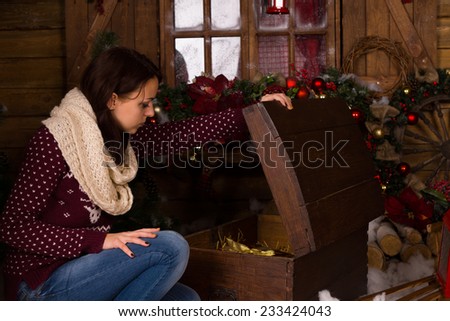 Sitting Young Woman in Trendy Winter Outfit Opening a Vintage Wooden Trunk inside the House with Christmas Decors.
