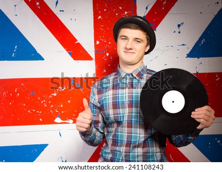 Close up Smiling Young Man in Checkered Long Sleeve Shirt and Hat Holding Vinyl Record Showing Thumbs Up While Looking at the Camera. Captured on Britain Flag Print Background.
