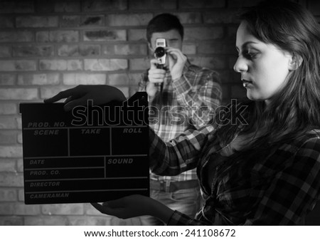 Serious Young Woman Holding Clapper Board with Young Male Photographer at the Back. Captured in Monochrome.