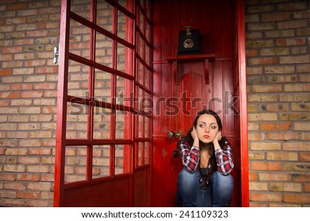 Troubled Young Woman with Hands Over Ears Crouching in Red Telephone Booth with Open Door