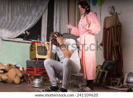 Woman in her dressing gown and slippers standing nagging her husband who is covering his ears in a squalid untidy living room