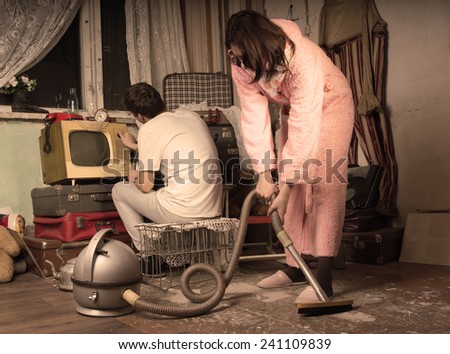 Retro housewife in her dressing gown and slippers cleaning a messy living room with a vintage vacuum cleaner while her husband watches television on an old TV set, aged style toning