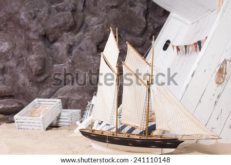 Toy yacht and wrecked wooden ship arranged in a marine or nautical still life an a sandy beach at the foot of a rocky cliff with wooden crates from the cargo in a conceptual image