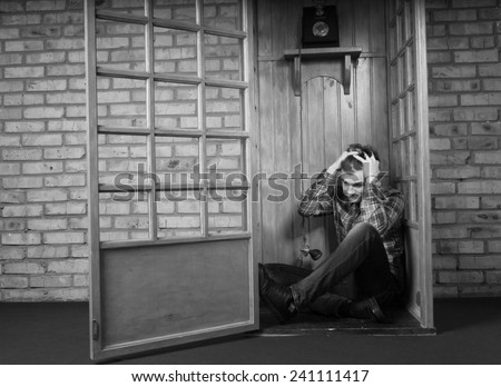 Troubled Man Sitting on Floor in Corner of Public Telephone Booth and Holding Head in Hands