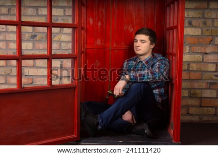 Bored young man waiting for a phone call on a public pay phone sitting on the floor of the booth with a listless expression