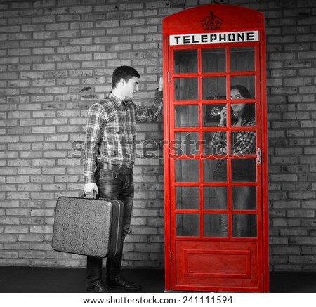 Young Handsome Man Holding Suitcase Knocking at the Telephone Booth with Woman Talking Inside. Captured with Gray Scale Effect.