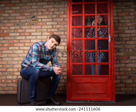 Man waiting for his girlfriend to stop talking on a public telephone sitting on his suitcase with a bored expression as she stands chatting to friends