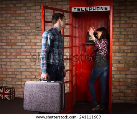 Woman refusing to listen to her husband as he implores her to get off the phone in a public telephone booth as he has a trip to make with his suitcase