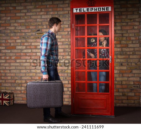 Man waiting with a suitcase as his wife makes a call on a telephone in a red British telephone booth
