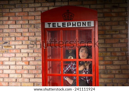 Young Woman Talking Someone Seriously in a Closed Red Telephone Booth with a Brick Wall Background.