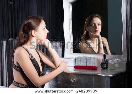 Pensive glamorous wistful young woman in evening wear sitting at her dressing table reflected in the mirror with a look of longing on her face