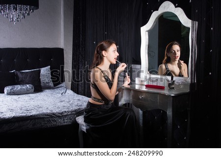 Glamorous young woman applying makeup in front of the mirror in an elegant bedroom in shadowed light